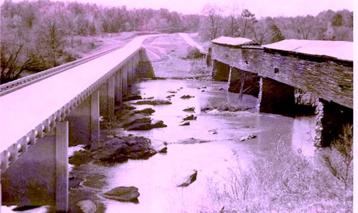 Black and white photo of a newly constructed bridge paralleling and older covered wooden bridge spanning a river.
