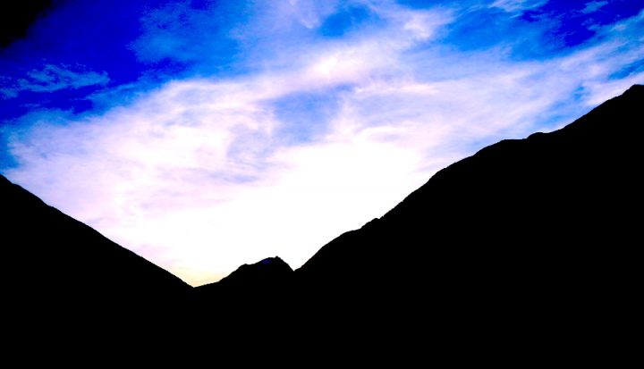 A blue sky with white clouds. The shadowy side of two mountains is in the foreground.