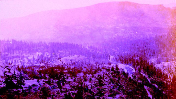 Old photograph of a hilly landscape with evergreen trees and snow. There is a valley running between the hills, and foggy weather conditions.