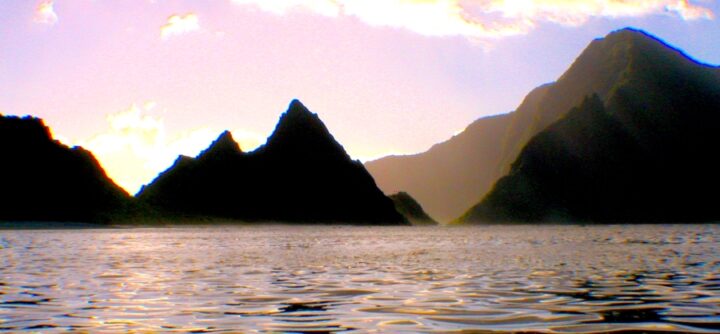 A body of water at dusk or dawn, with mountains jetting out of the water, forming islands or peninsulas. The sky is light purple and there is a light mist.