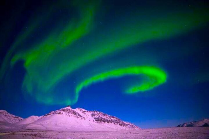 Florescent green aurora bands in a dark blue sky above a melting snowy polar landscape.