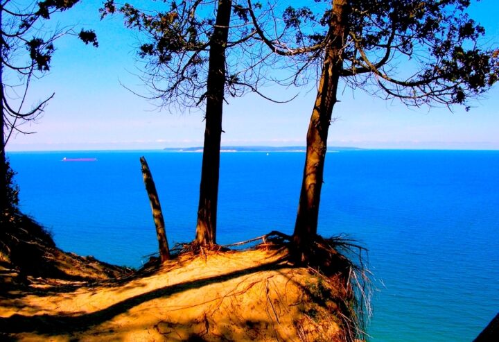 And sand-covered cliff edge with deciduous trees. The cliff is overlooking a large lake that is deep blue. The sky in the background is blue. There is some haze in the distance.