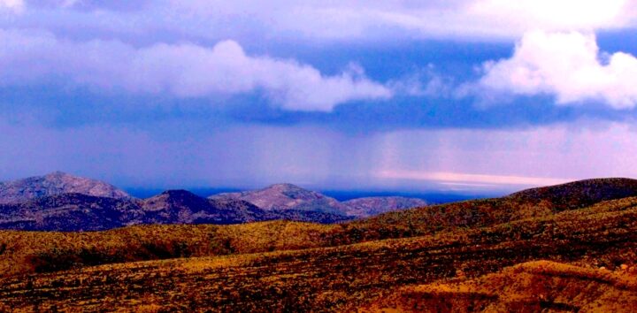 A desert landscape featuring a distance rainstorm. In the foreground are short, orange hills without vegetation. Behind that are taller hills that appear blue, and are also without vegetation. Behind that is a blue cloudy sky, with two curtains of rain reaching the landscape.
