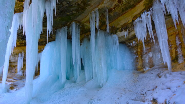 Icicles hanging from below an escarpment. The icycles are robust and light blue. There is white ice on the ground.