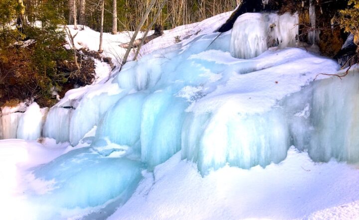 A fully frozen cascading waterfall covered mostly in snow. The waterway ice is blue. Trees and bushes surround the waterway.