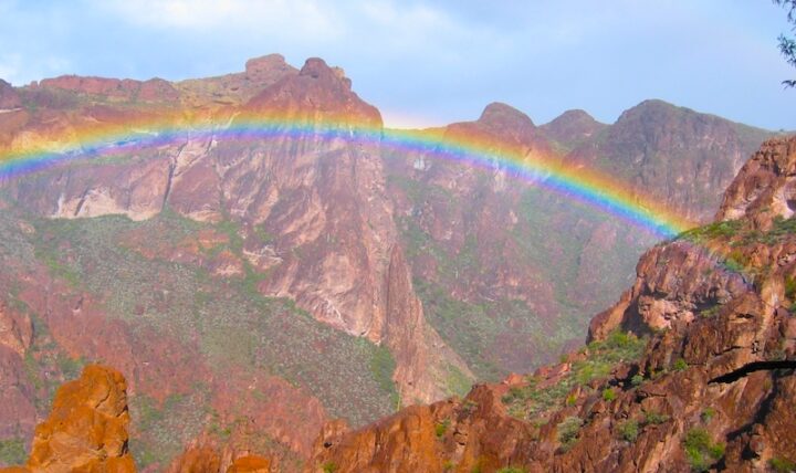 Desert mountain valley with a rainbow arcing below a mostly clear blue sky. Light rain falls on the dry brown ground where scattered desert plants grow.