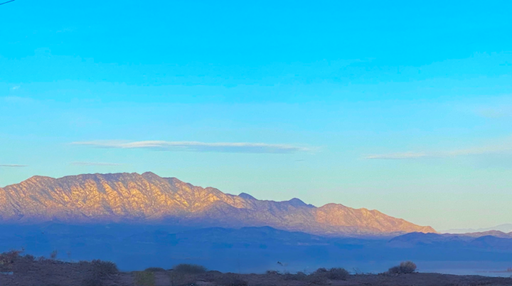 Sunrise light illuminates the mountain peak while the base remains in shadow, with dark blue tones. The sky above is blue with a few scattered clouds.